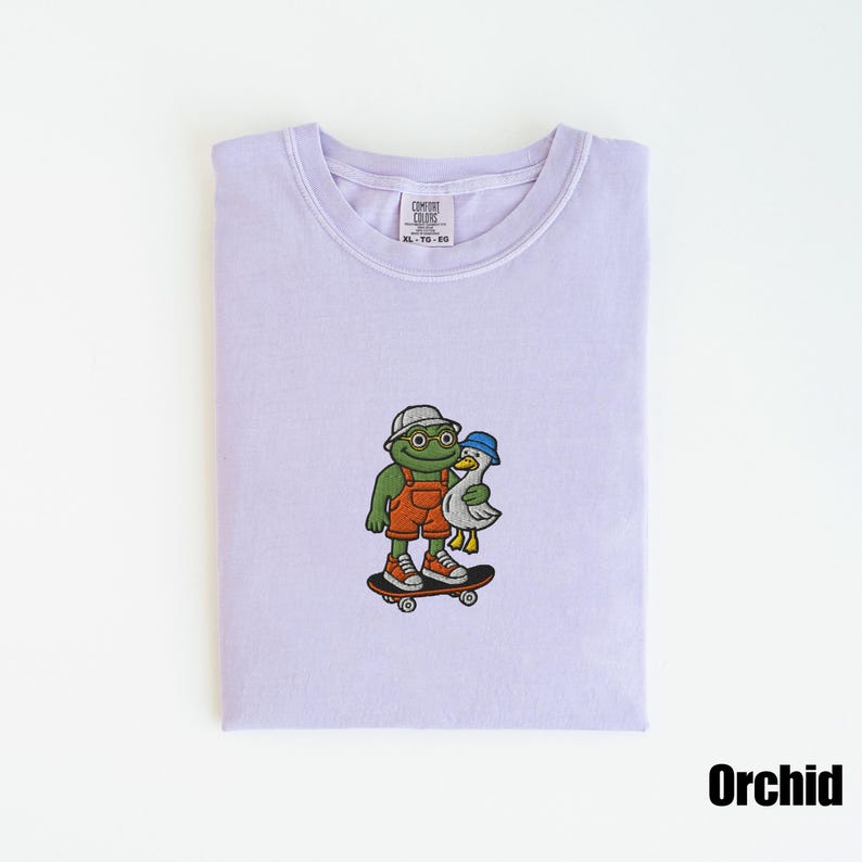 May include: Orchid-colored t-shirt with an embroidered design of a frog wearing overalls, a hat, and sunglasses, skateboarding with a duck. The shirt has a crew neck and a tag that reads "Comfort Colors". The word "Orchid" is printed at the bottom.