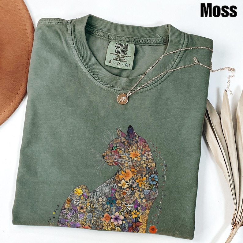 May include: A green t-shirt with a floral cat design. The shirt has the text "Comfort Colors" and "S - P - CH" printed on the tag. The shirt is laid flat on a surface.