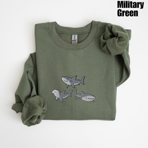 May include: Military green sweatshirt with embroidered shark designs. The crew neck sweatshirt features three different shark illustrations in shades of gray and white. The text "Military Green" is in the upper right corner.