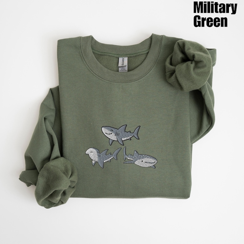 May include: Military green sweatshirt with embroidered shark designs. The crew neck sweatshirt features three different shark illustrations in shades of gray and white. The text "Military Green" is in the upper right corner.