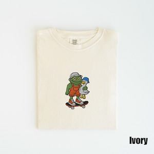 May include: Ivory-colored t-shirt with a crew neck. The shirt features an embroidered design of a green frog wearing a hat and overalls, skateboarding with a duck. The word "Ivory" is printed at the bottom right.