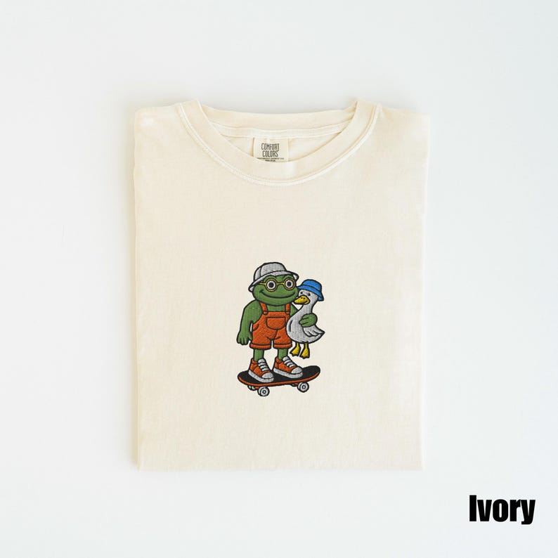 May include: Ivory-colored t-shirt with a crew neck. The shirt features an embroidered design of a green frog wearing a hat and overalls, skateboarding with a duck. The word "Ivory" is printed at the bottom right.