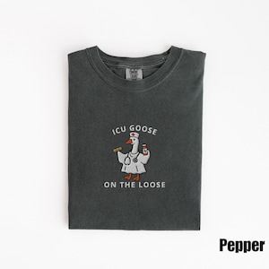May include: A dark gray t-shirt with the text "ICU GOOSE ON THE LOOSE" and a cartoon goose wearing a doctor's coat and holding a stethoscope and medicine bottle. The shirt is folded neatly on a white surface.