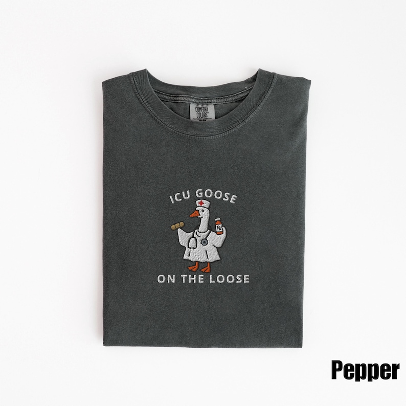 May include: A dark gray t-shirt with the text "ICU GOOSE ON THE LOOSE" and a cartoon goose wearing a doctor's coat and holding a stethoscope and medicine bottle. The shirt is folded neatly on a white surface.