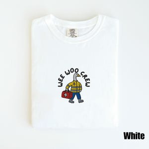 May include: White t-shirt with a crew neck featuring a cartoon duck in a yellow jacket, blue pants, and a red medical bag. The text "WEE WOO CREW" is above the duck. The word "White" is at the bottom right.