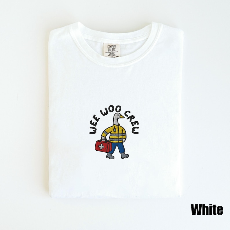 May include: White t-shirt with a crew neck featuring a cartoon duck in a yellow jacket, blue pants, and a red medical bag. The text "WEE WOO CREW" is above the duck. The word "White" is at the bottom right.