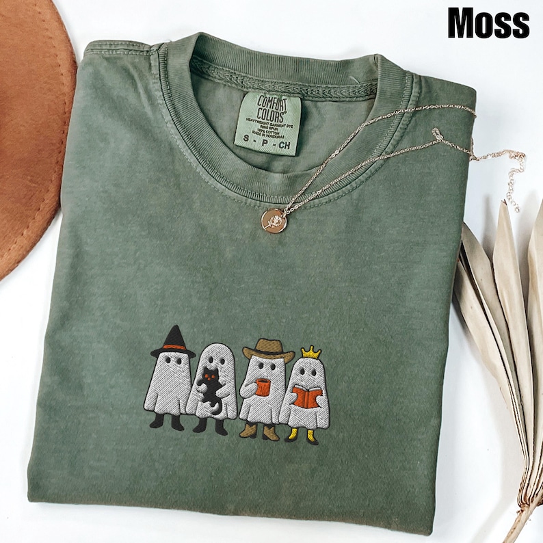May include: A moss green t-shirt with an embroidered design of four cartoon ghosts, each with a unique accessory. The ghosts are wearing a witch hat, holding a black cat, a cowboy hat, and a crown. A gold necklace with a pendant is draped on the shirt.