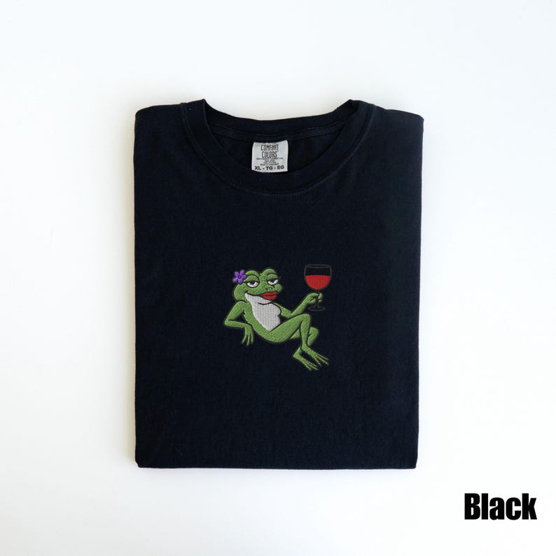 Pu&ograve; includere: T-shirt nero con un disegno ricamato di una rana verde con un fiore viola, rossetto rosso e un bicchiere di vino rosso. La maglietta ha un girocollo e maniche corte. Il testo "Black" &egrave; in basso a destra.
