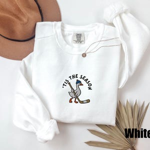May include: White crew neck sweatshirt with the phrase "'Tis the Season" and a cartoon goose wearing a blue hat and holding a hockey stick. The sweatshirt has long sleeves and a tag that says "Comfort Colors". A gold necklace is also visible.