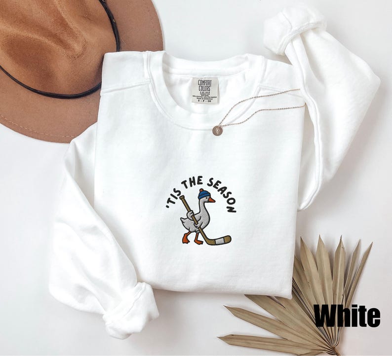 May include: White crew neck sweatshirt with the phrase "'Tis the Season" and a cartoon goose wearing a blue hat and holding a hockey stick. The sweatshirt has long sleeves and a tag that says "Comfort Colors". A gold necklace is also visible.