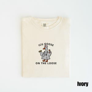 May include: Ivory-colored t-shirt with a crew neck. The shirt features an embroidered cartoon goose wearing a doctor's coat, stethoscope, and hat, holding a syringe and medicine bottle. Text reads "ICU GOOSE ON THE LOOSE".