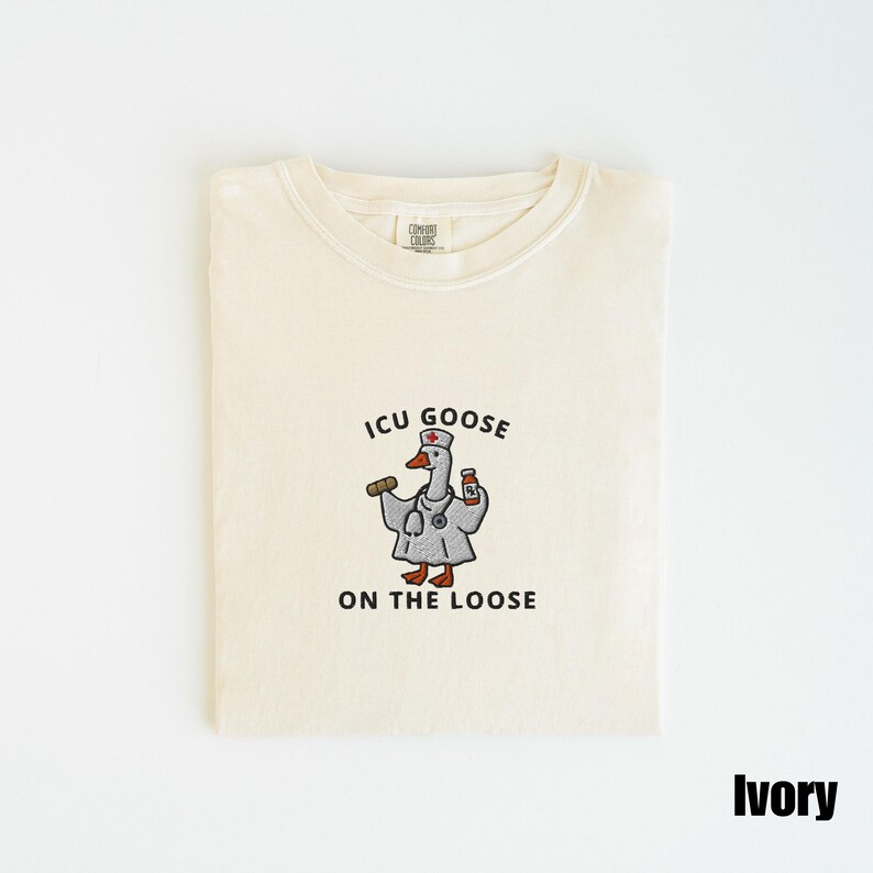 May include: Ivory-colored t-shirt with a crew neck. The shirt features an embroidered cartoon goose wearing a doctor's coat, stethoscope, and hat, holding a syringe and medicine bottle. Text reads "ICU GOOSE ON THE LOOSE".