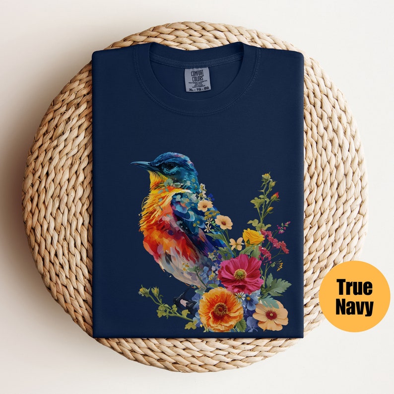 May include: A navy blue t-shirt featuring a colourful bird and floral design. The bird is painted in shades of blue, yellow, orange, and red. The flowers are in shades of pink, yellow, and orange.