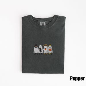 May include: Dark gray t-shirt with an embroidered design of four cartoon ghosts, each with a unique accessory: a witch hat, a black cat, a cowboy hat, and a crown. The word "Pepper" is in the bottom right corner.