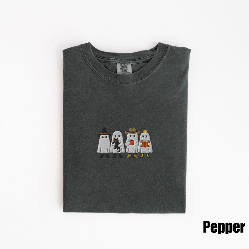 May include: Dark gray t-shirt with an embroidered design of four cartoon ghosts, each with a unique accessory: a witch hat, a black cat, a cowboy hat, and a crown. The word "Pepper" is in the bottom right corner.
