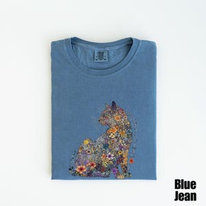 May include: A blue jean t-shirt with a floral cat design. The cat is made up of colorful flowers and is sitting in a relaxed pose. The t-shirt is folded and the brand tag is visible.