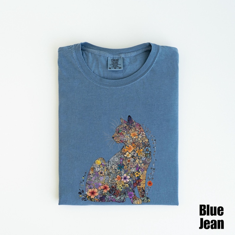 May include: A blue jean t-shirt with a floral cat design. The cat is made up of colorful flowers and is sitting in a relaxed pose. The t-shirt is folded and the brand tag is visible.
