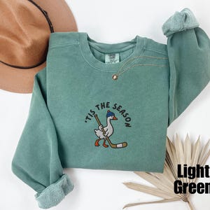 May include: Light green sweatshirt with the phrase "'Tis the Season" embroidered above a cartoon goose wearing a hat and holding a hockey stick. The sweatshirt has a crew neck and long sleeves.