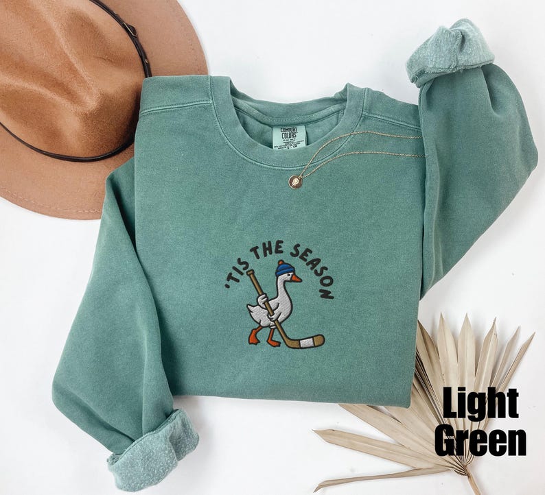 May include: Light green sweatshirt with the phrase "'Tis the Season" embroidered above a cartoon goose wearing a hat and holding a hockey stick. The sweatshirt has a crew neck and long sleeves.