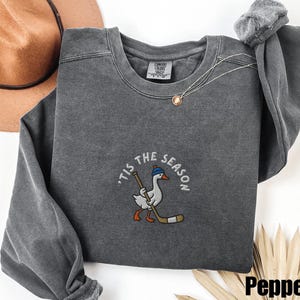 May include: A gray crewneck sweatshirt with the embroidered phrase "'Tis the Season" and a cartoon goose wearing a hat and holding a hockey stick. The sweatshirt has a relaxed fit and a vintage wash.