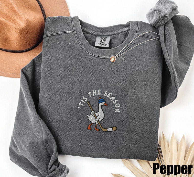 May include: A gray crewneck sweatshirt with the embroidered phrase "'Tis the Season" and a cartoon goose wearing a hat and holding a hockey stick. The sweatshirt has a relaxed fit and a vintage wash.