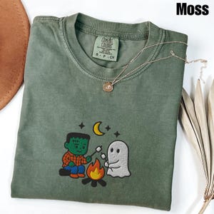 May include: A moss green Comfort Colors t-shirt with an embroidered design of a Frankenstein-like character and a ghost roasting marshmallows over a campfire. A gold necklace with a pendant is draped over the shirt.