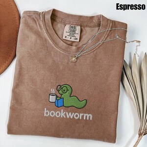 May include: Espresso-colored t-shirt with a crew neck. The shirt features an embroidered green bookworm wearing glasses, holding a coffee cup and a blue book. The word "bookworm" is embroidered below the graphic. A silver necklace with a pendant is draped across the shirt.