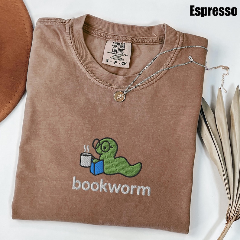 May include: Espresso-colored t-shirt with a crew neck. The shirt features an embroidered green bookworm wearing glasses, holding a coffee cup and a blue book. The word "bookworm" is embroidered below the graphic. A silver necklace with a pendant is draped across the shirt.