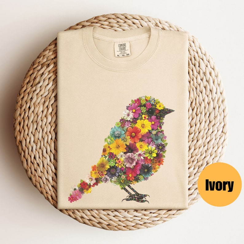 May include: A beige t-shirt with a floral bird design. The bird is made up of colorful flowers and is facing right. The shirt is on a woven wicker surface. The text "COMFORT COLORS" is on the tag. The color of the shirt is ivory.