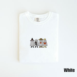 May include: White t-shirt featuring an embroidered design of four ghosts dressed in costumes. One ghost wears a witch hat, another holds a black cat, a third holds a coffee cup, and the last reads a book. The word "White" is printed at the bottom.
