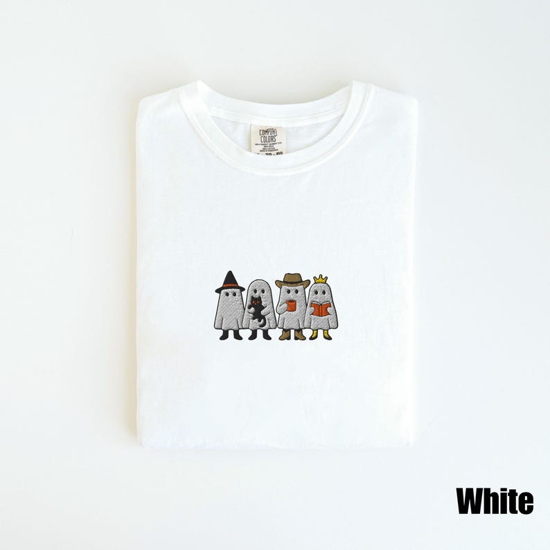 May include: White t-shirt featuring an embroidered design of four ghosts dressed in costumes. One ghost wears a witch hat, another holds a black cat, a third holds a coffee cup, and the last reads a book. The word "White" is printed at the bottom.