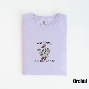 May include: A light purple t-shirt with the text "ICU GOOSE ON THE LOOSE" and a cartoon goose wearing a doctor's coat and stethoscope. The goose is holding a pill and a medicine bottle. The shirt is labeled "Orchid".