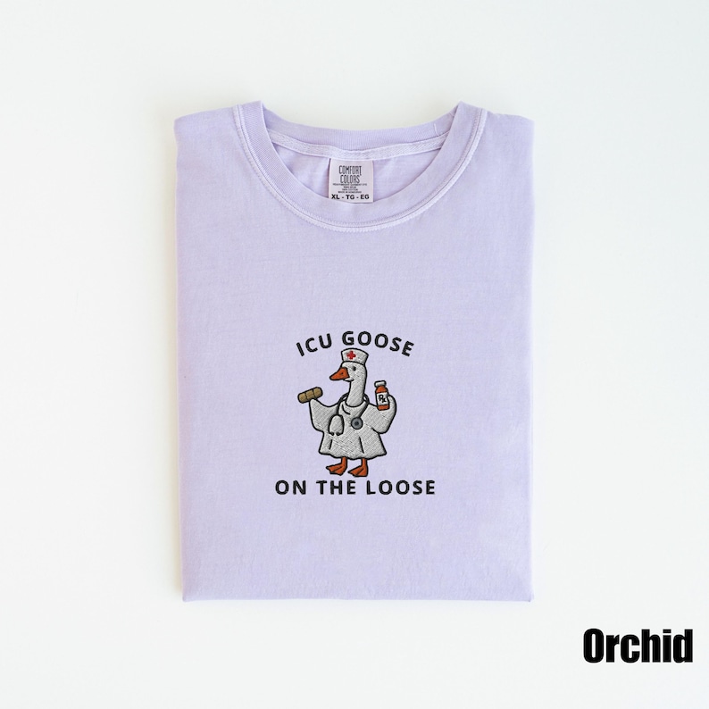 May include: A light purple t-shirt with the text "ICU GOOSE ON THE LOOSE" and a cartoon goose wearing a doctor's coat and stethoscope. The goose is holding a pill and a medicine bottle. The shirt is labeled "Orchid".
