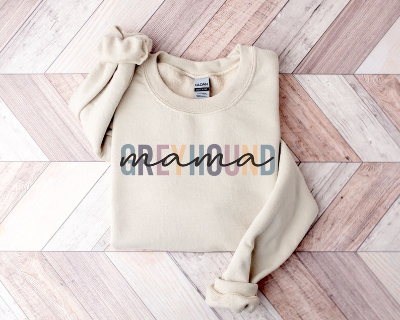 May include: A beige sweatshirt with the text "Greyhound mama" in colorful letters. The letters are in shades of pink, yellow, blue, and gray.