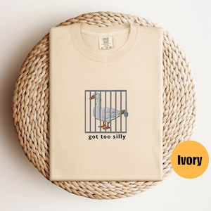 May include: A beige t-shirt with a cartoon goose in a cage and the text "got too silly". The shirt is on a woven surface. The color of the shirt is ivory.