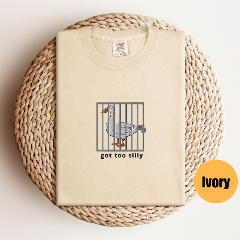 May include: A beige t-shirt with a cartoon goose in a cage and the text "got too silly". The shirt is on a woven surface. The color of the shirt is ivory.