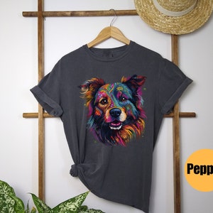 May include: A dark grey t-shirt featuring a colourful, psychedelic illustration of a dog's face. The dog has a pink nose and blue eyes. The text "Pepper" is printed on a yellow circle in the bottom right corner of the image.
