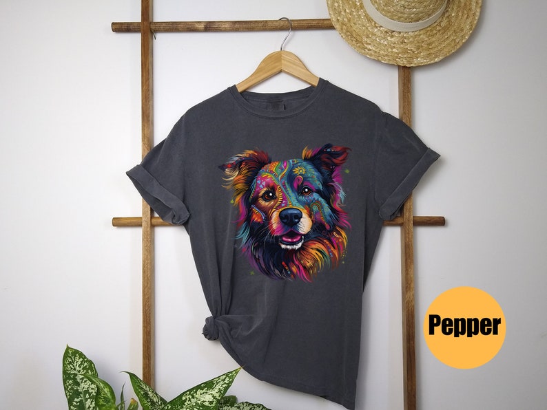 May include: A dark grey t-shirt featuring a colourful, psychedelic illustration of a dog's face. The dog has a pink nose and blue eyes. The text "Pepper" is printed on a yellow circle in the bottom right corner of the image.