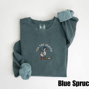 May include: Blue Spruce sweatshirt with a crew neck and long sleeves. The front features an embroidered design of a goose wearing a hat and holding a hockey stick, with the text "'Tis the Season" around it. The text "Blue Spruce" is at the bottom.