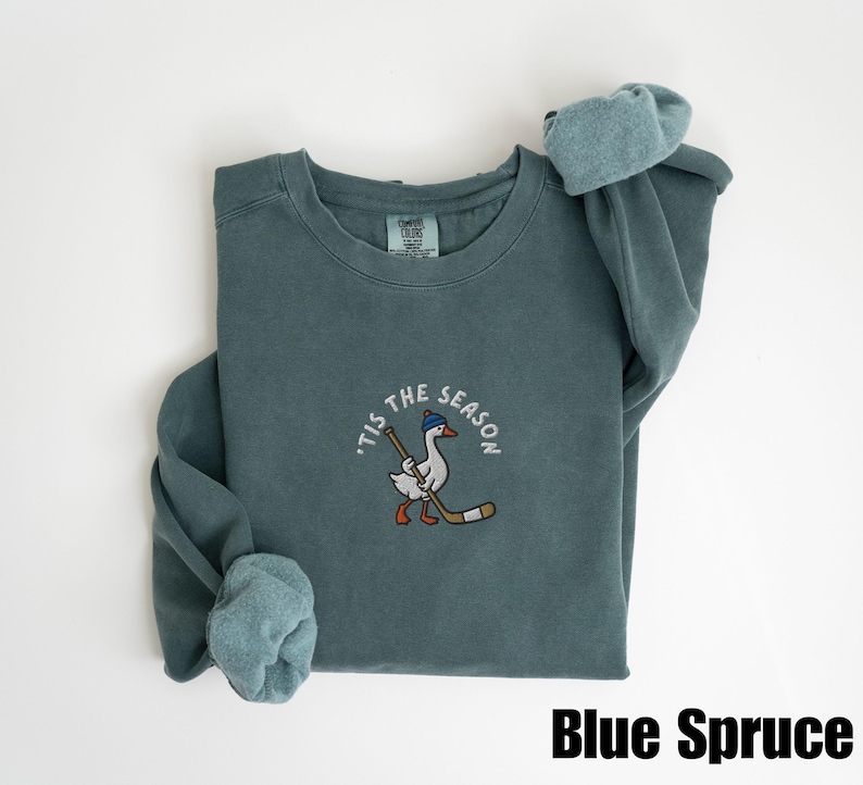 May include: Blue Spruce sweatshirt with a crew neck and long sleeves. The front features an embroidered design of a goose wearing a hat and holding a hockey stick, with the text "'Tis the Season" around it. The text "Blue Spruce" is at the bottom.