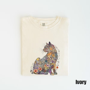 May include: Ivory t-shirt with a floral cat design. The cat is made up of colorful flowers and leaves. The shirt has a crew neck and short sleeves. The text "COMFORT COLORS" is visible on the tag.
