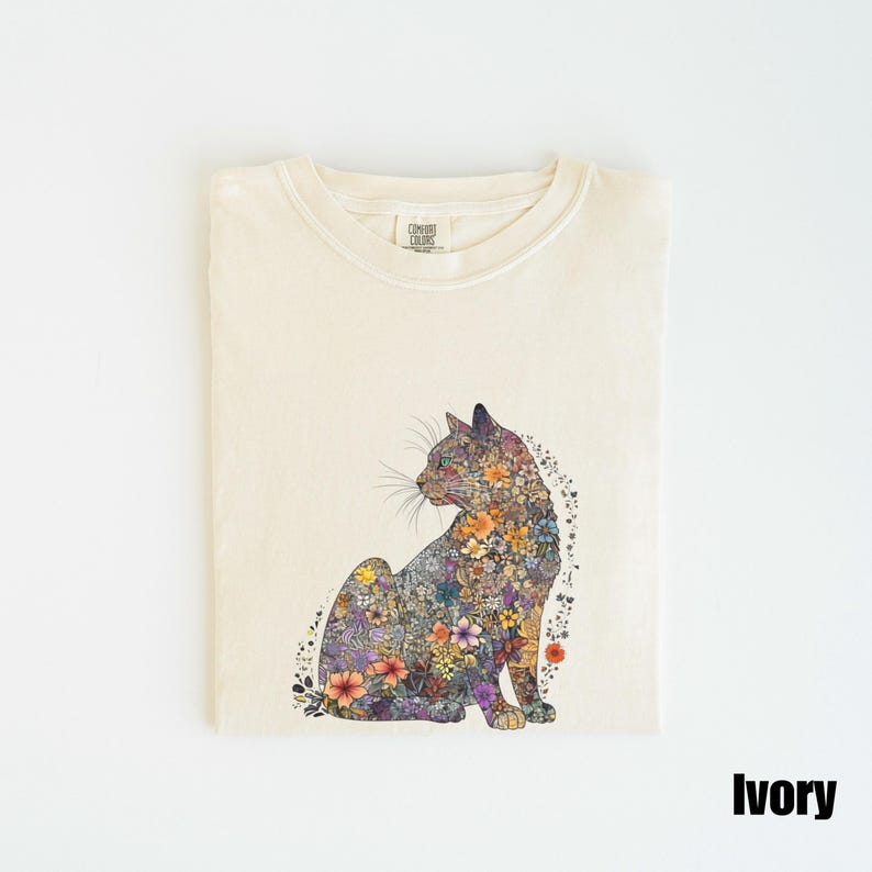 May include: Ivory t-shirt with a floral cat design. The cat is made up of colorful flowers and leaves. The shirt has a crew neck and short sleeves. The text "COMFORT COLORS" is visible on the tag.