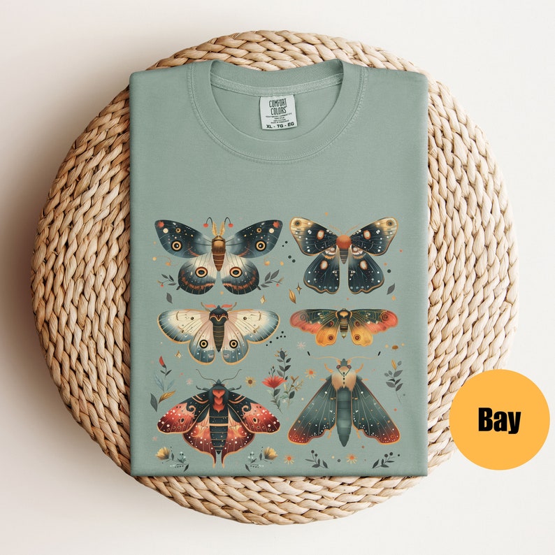 Puede incluir: Una camiseta verde claro con un estampado de seis polillas coloridas con detalles intrincados. Las polillas est&aacute;n rodeadas de peque&ntilde;as flores y hojas. La camiseta tiene una etiqueta que dice "COMFORT COLORS".