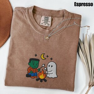 May include: Espresso-colored t-shirt with an embroidered design featuring a Frankenstein-like character and a ghost roasting marshmallows over a campfire. The shirt has a crew neck and a Comfort Colors tag. A silver necklace with a pendant is draped across the shirt.
