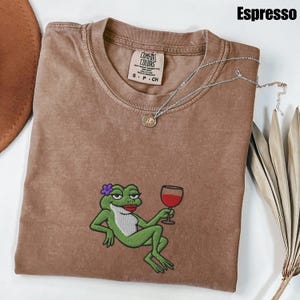 Pu&ograve; includere: T-shirt color espresso con un ricamo di una rana verde che tiene in mano un bicchiere di vino rosso. La rana indossa un fiore viola e rossetto rosso. Una collana d'argento con un piccolo ciondolo &egrave; appoggiata sulla maglietta. La maglietta ha un'etichetta Comfort Colors.