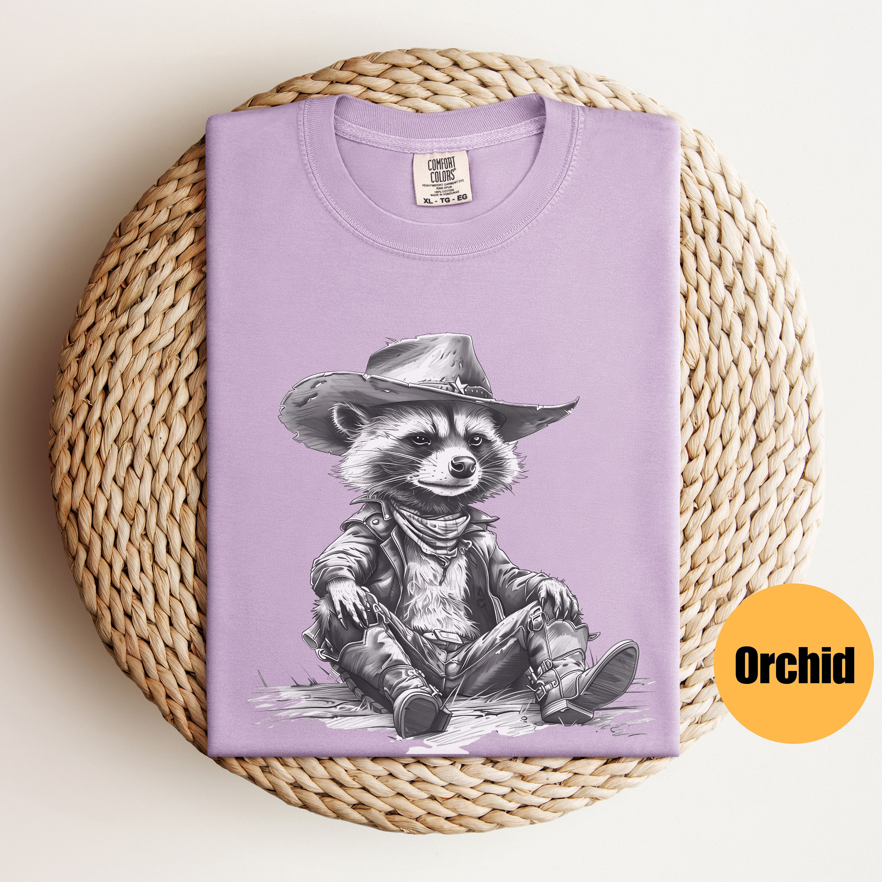 May include: A lavender t-shirt with a black and white illustration of a raccoon wearing a cowboy hat and a leather jacket. The raccoon is sitting with its legs crossed and wearing cowboy boots. The text "COMFORT COLORS" is visible on the tag.