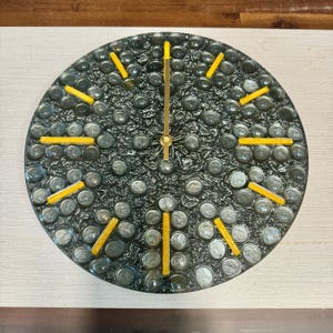 May include: A round wall clock with a textured, dark gray face. The clock face is decorated with small, round, gray stones and yellow markers. The clock hands are gold-colored.
