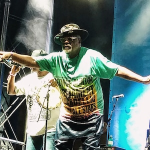 Può includere: Un uomo che indossa una camicia verde, gialla e nera con il testo "CONNECTION PRESENTS" e "ALL STARS" su di essa. Indossa un cappello nero e tiene un microfono in mano. È in piedi su un palco con uno sfondo blu.