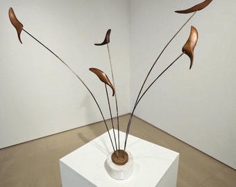 May include: Abstract sculpture featuring six brown, leaf-shaped elements attached to thin, curved metal rods. The rods extend from a circular base atop a white cube. The sculpture is set against a plain white background, creating a minimalist aesthetic.