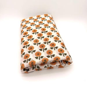May include: A folded piece of white fabric featuring a repeating pattern of orange flowers with dark green stems and leaves. The textile has a textured, crinkled appearance and is arranged in a rectangular stack against a plain white background.